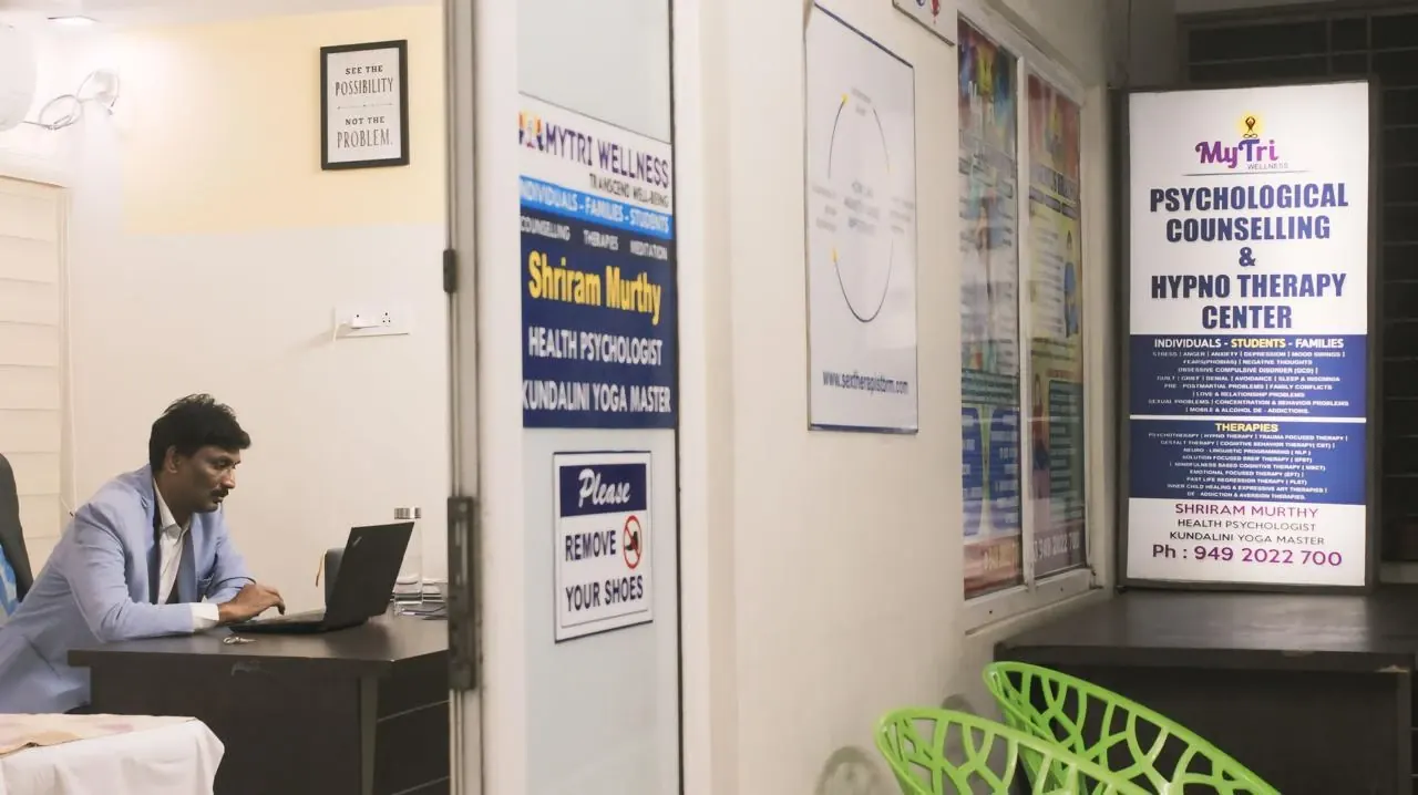 Dr. Shriram Murthy at his desk at MyTri Wellness, Maharanipeta, beside the centre's counselling and hypnotherapy sign