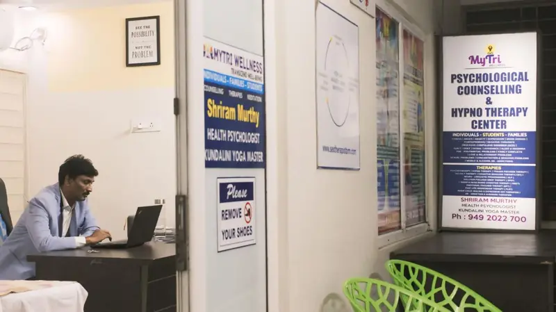 Dr. Shriram Murthy at his desk at MyTri Wellness, Maharanipeta, beside the centre's counselling and hypnotherapy sign